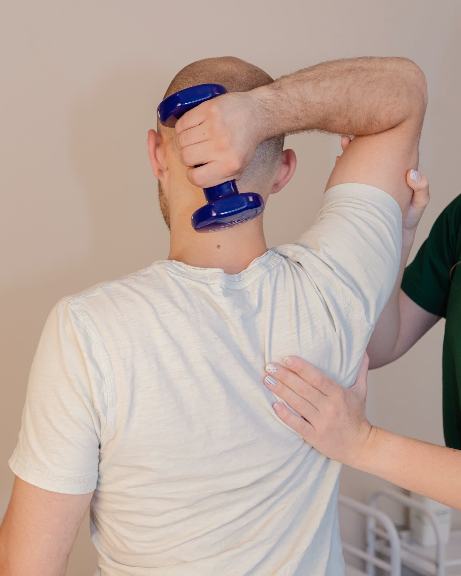 Кинезитерапевт води упражнение за рамо с дъмбел: клиентът държи дъмбела зад главата, терапевтът придържа лакътя и лопатката